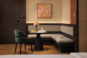 Habitación de hotel moderna con puerta de madera oscura, sillón tapizado en verde, una mesa redonda con un jarrón de flores, sofá gris con un cojín y un cuadro abstracto colorido en la pared.