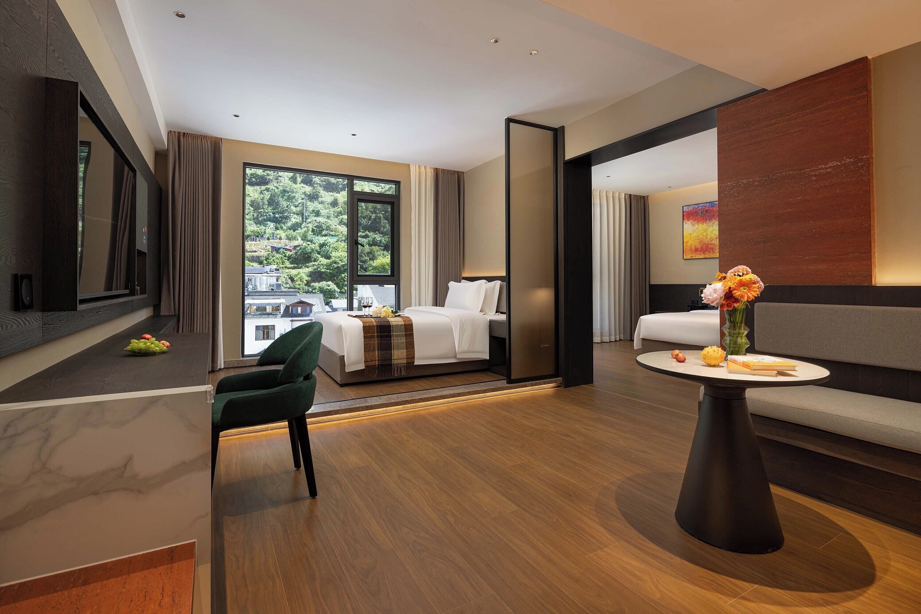 A modern hotel room with a bed, a sofa, a dining table, and a view of greenery outside.
