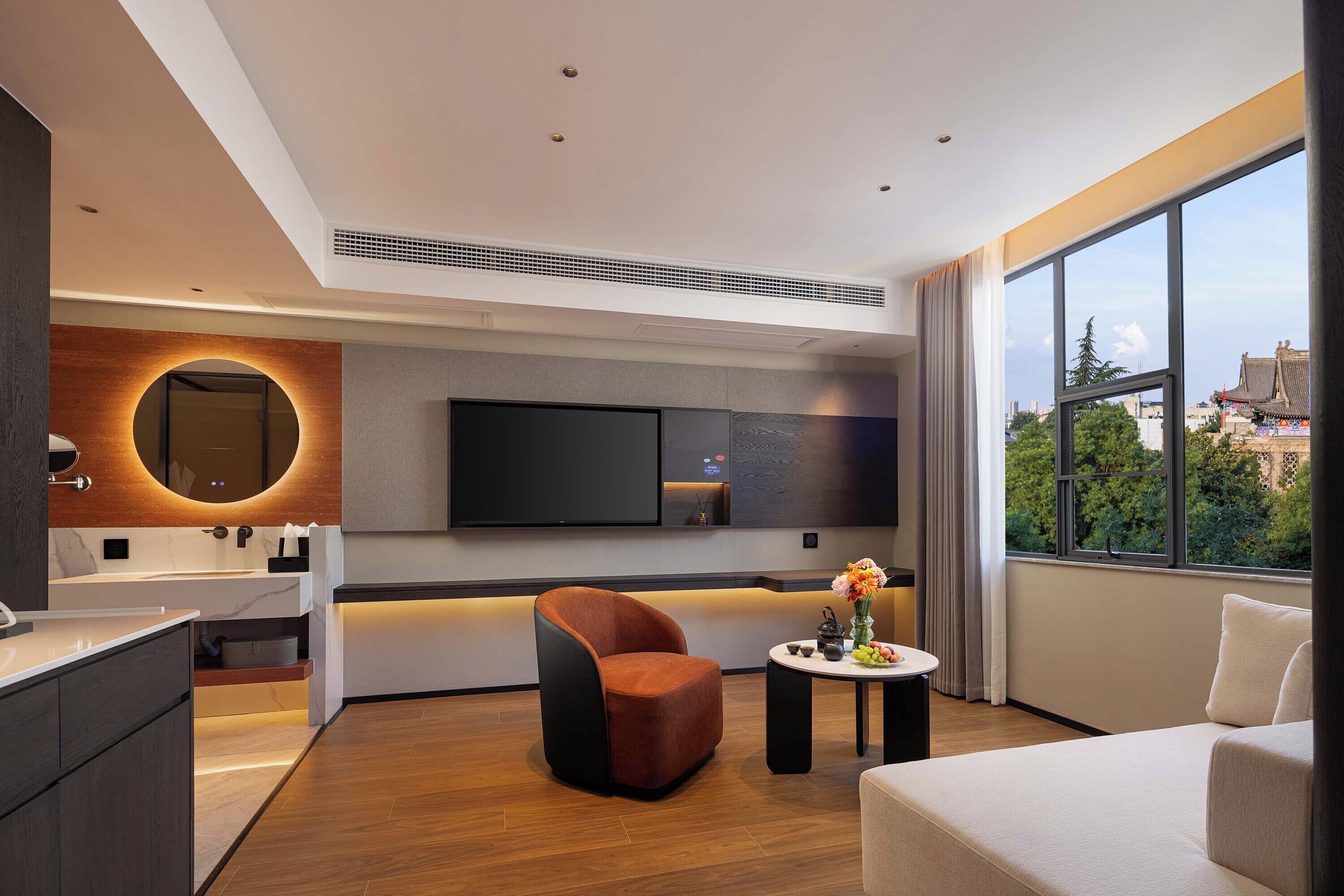 A modern hotel room with a large window, a flat-screen TV, a round mirror, a sofa, and a small table with a vase of flowers.