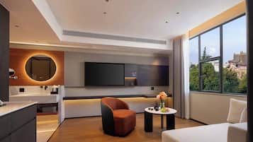 A modern hotel room with a large window, a flat-screen TV, a round mirror, a sofa, and a small table with a vase of flowers.