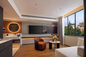 Quarto de hotel moderno com uma grande janela, uma TV de tela plana, um espelho redondo, um sofá e uma pequena mesa com um vaso de flores.