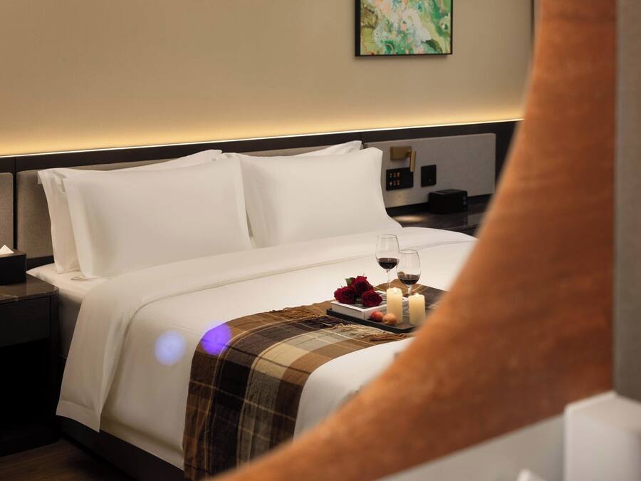 Habitación de hotel con una cama bien hecha, una mesita de noche con lámpara y una bandeja con copas de vino y flores.