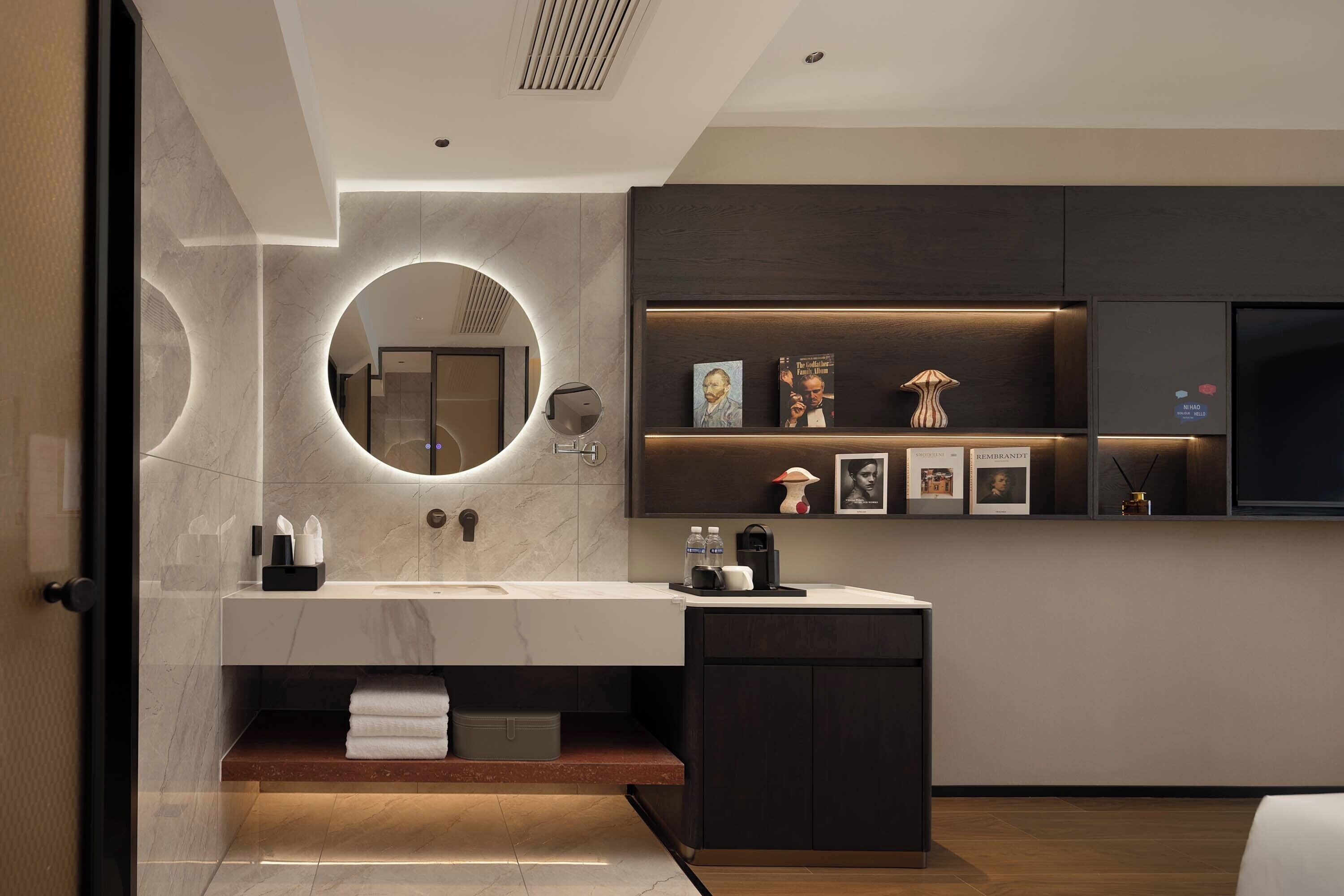 Une salle de bain moderne avec un meuble-lavabo en marbre, un miroir rond et une étagère où sont posés des livres et des objets décoratifs.