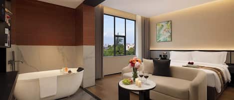 A modern hotel room with a bathtub, a sofa, a bedside table with a vase of flowers, and a large window offering a city view.
