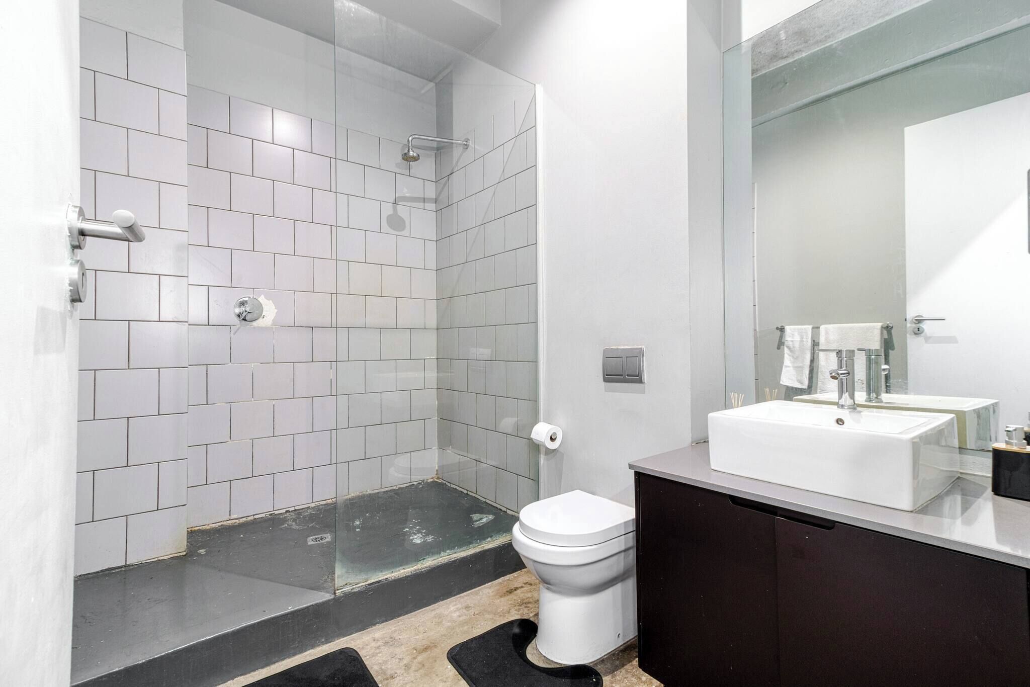 A modern bathroom with white tiled walls, a glass shower enclosure, a white toilet, and a dark-toned vanity with a mirror.