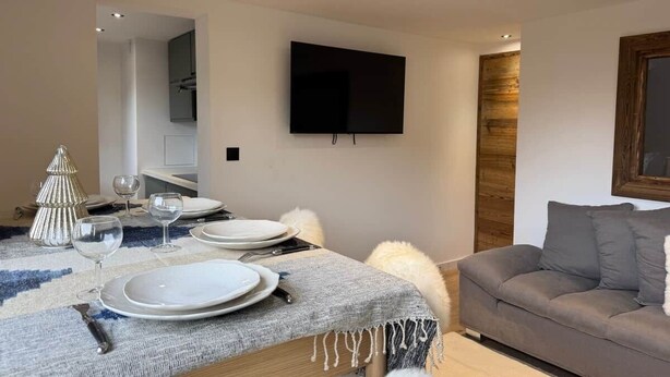 Ein moderner Essbereich mit einem Tisch für vier Personen, einem grauen Sofa und einem an der Wand montierten Fernseher.