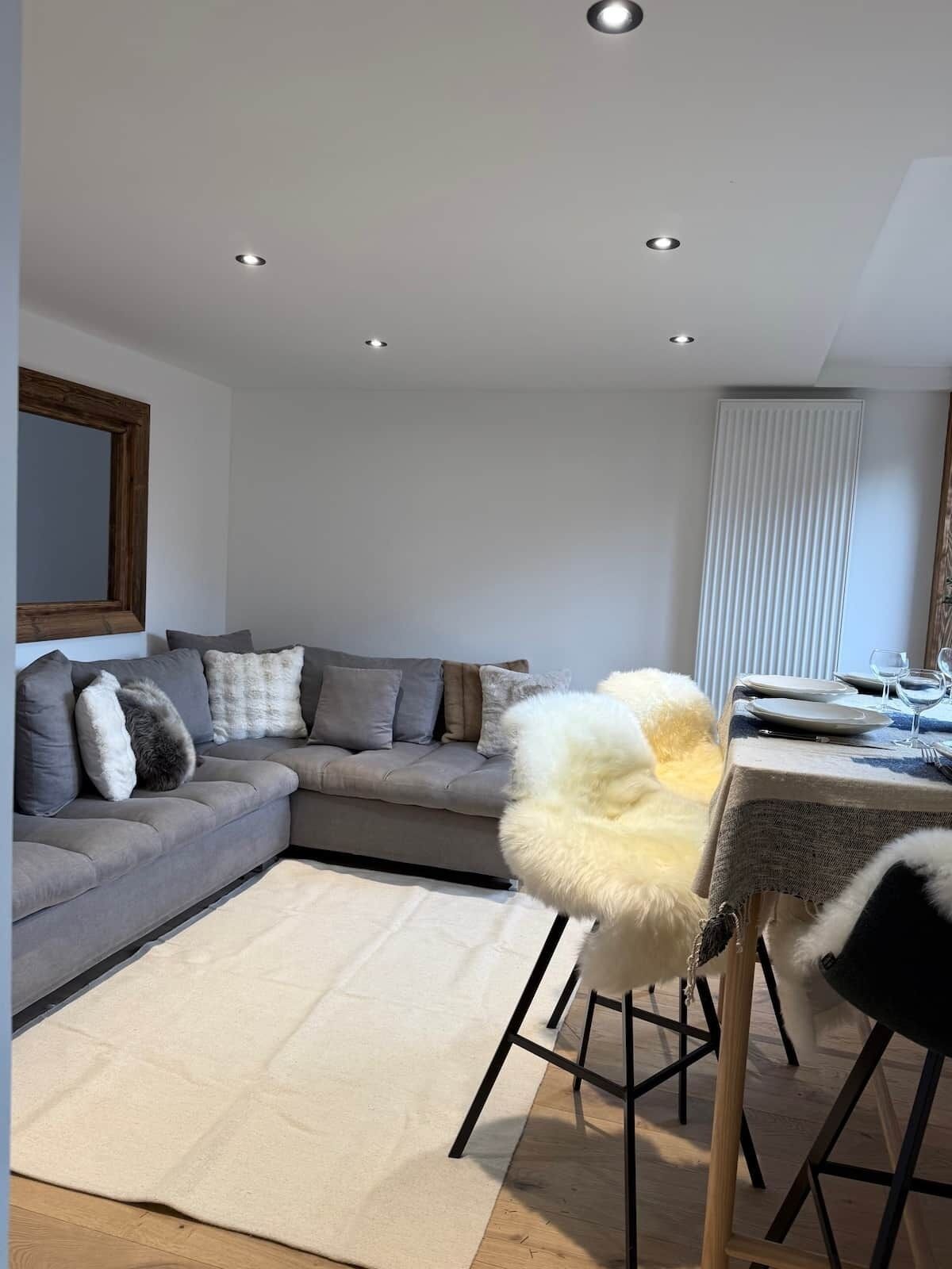 Ein modernes Wohnzimmer mit einem grauen Ecksofa, einem weißen, flauschigen Sessel, einem Esstisch mit Stühlen und Einbauleuchten.