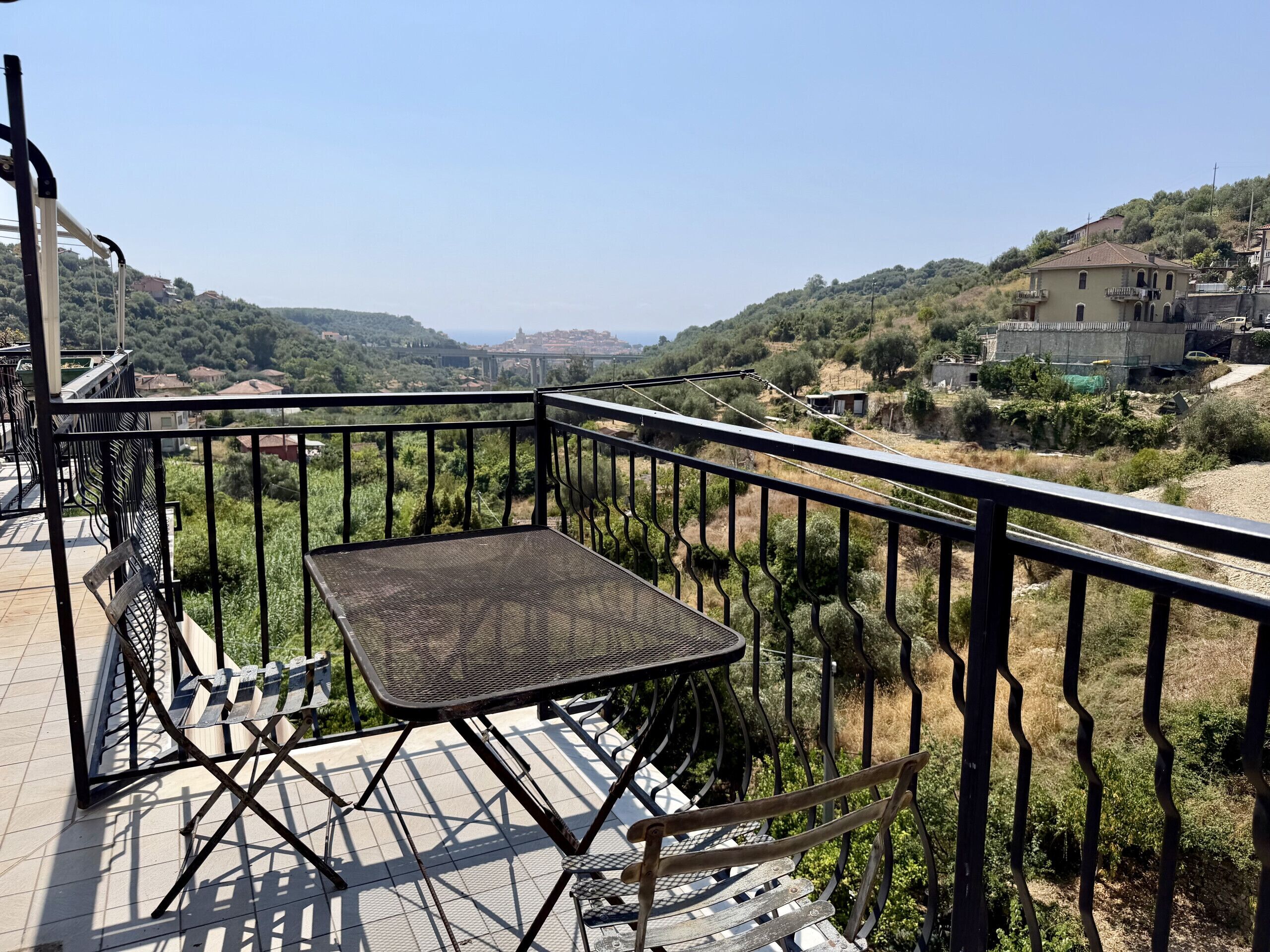A balcony with a metal table and chairs, overlooking a hilly landscape with buildings.