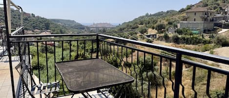 A balcony with a metal table and chairs, overlooking a hilly landscape with buildings.