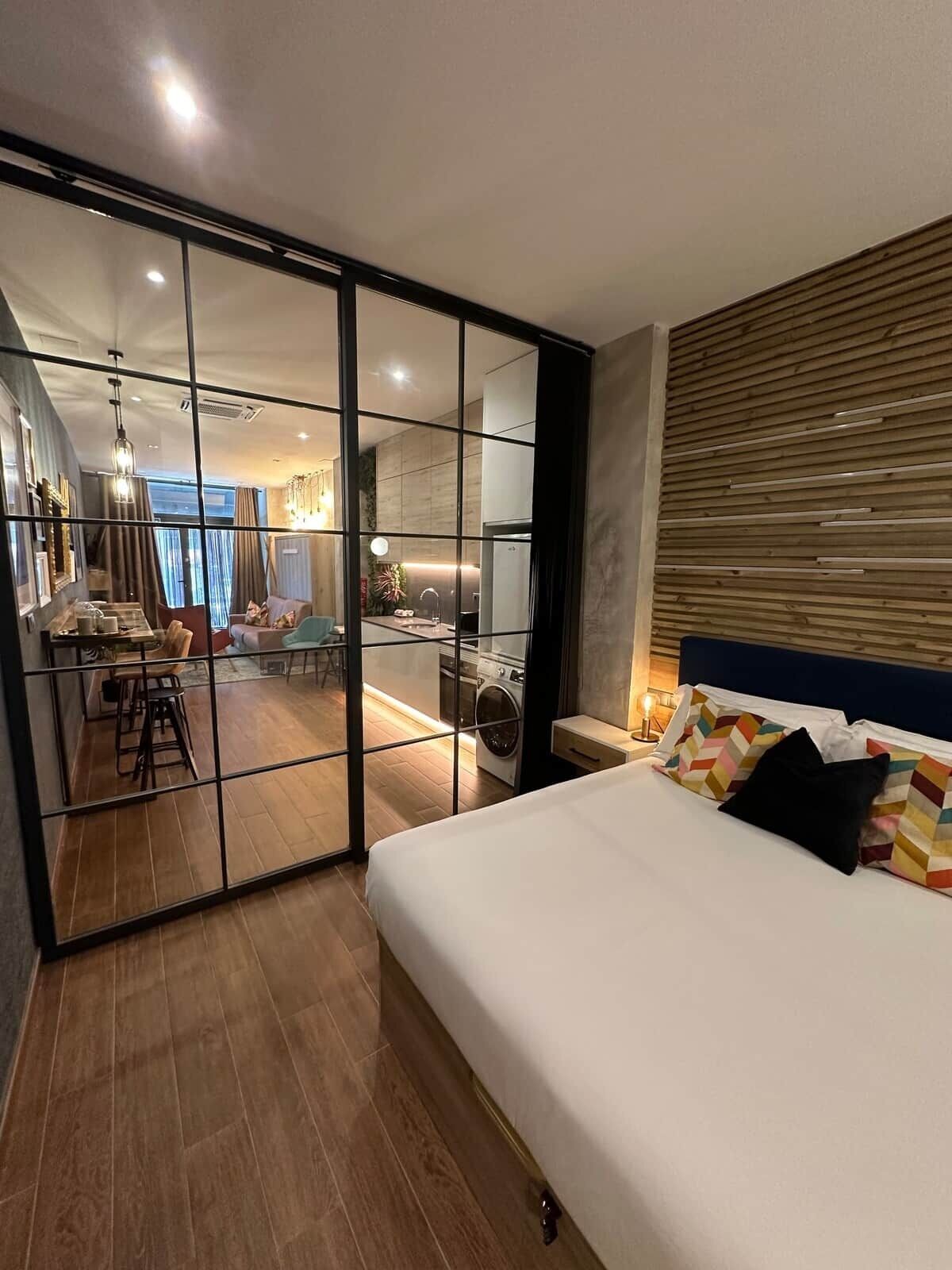 Une chambre d’hôtel moderne dotée d’un grand lit, d’une tête de lit en bois, d’une porte-fenêtre coulissante donnant sur un coin repas et d’un espace buanderie visible.