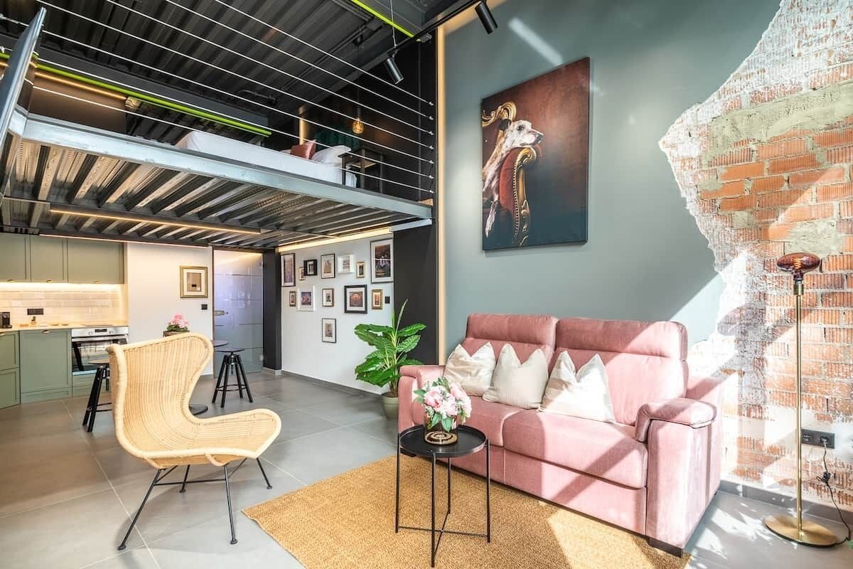 A modern living space with a loft area, a pink sofa, a wicker chair, and a large artwork on the wall.