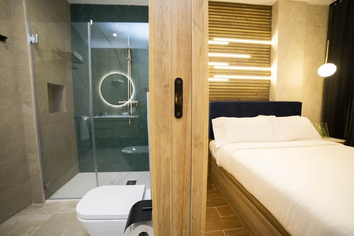 A hotel room with a glass-enclosed shower, a bed with white bedding, and a wooden headboard.
