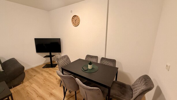 Een eethoek met een rechthoekige tafel, zes stoelen, een televisie op een dressoir, en een klok aan de muur.