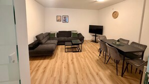 Een woonkamer met een modulaire bank, een eettafel met stoelen, een flatscreen televisie en een klok aan de muur.