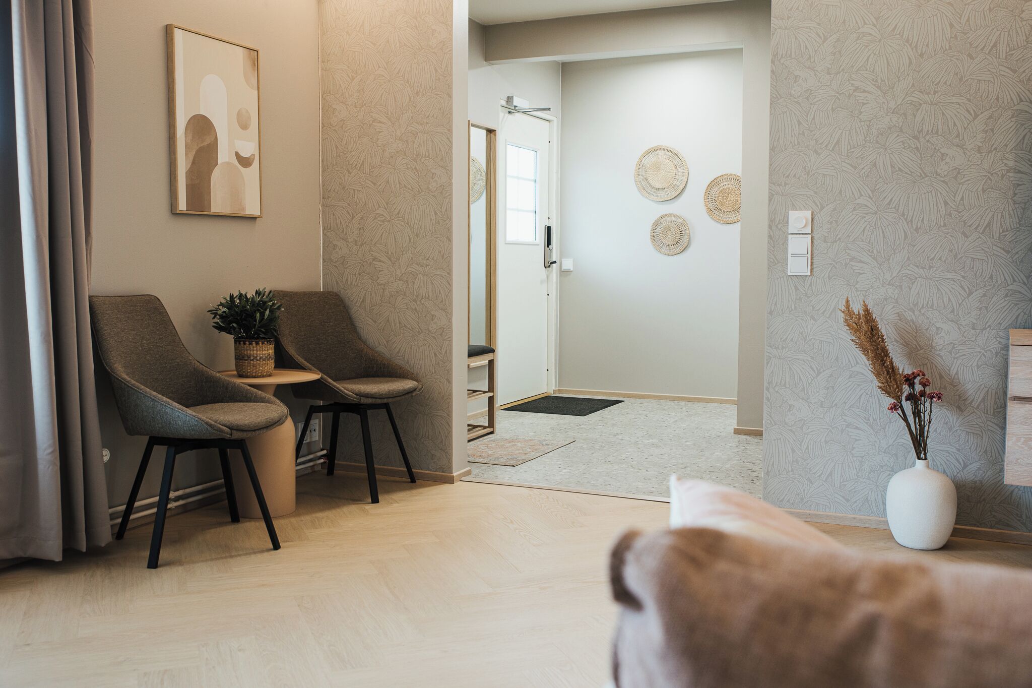 A modern living room with two armchairs, a coffee table, a vase with dried flowers, and a door leading to a hallway.