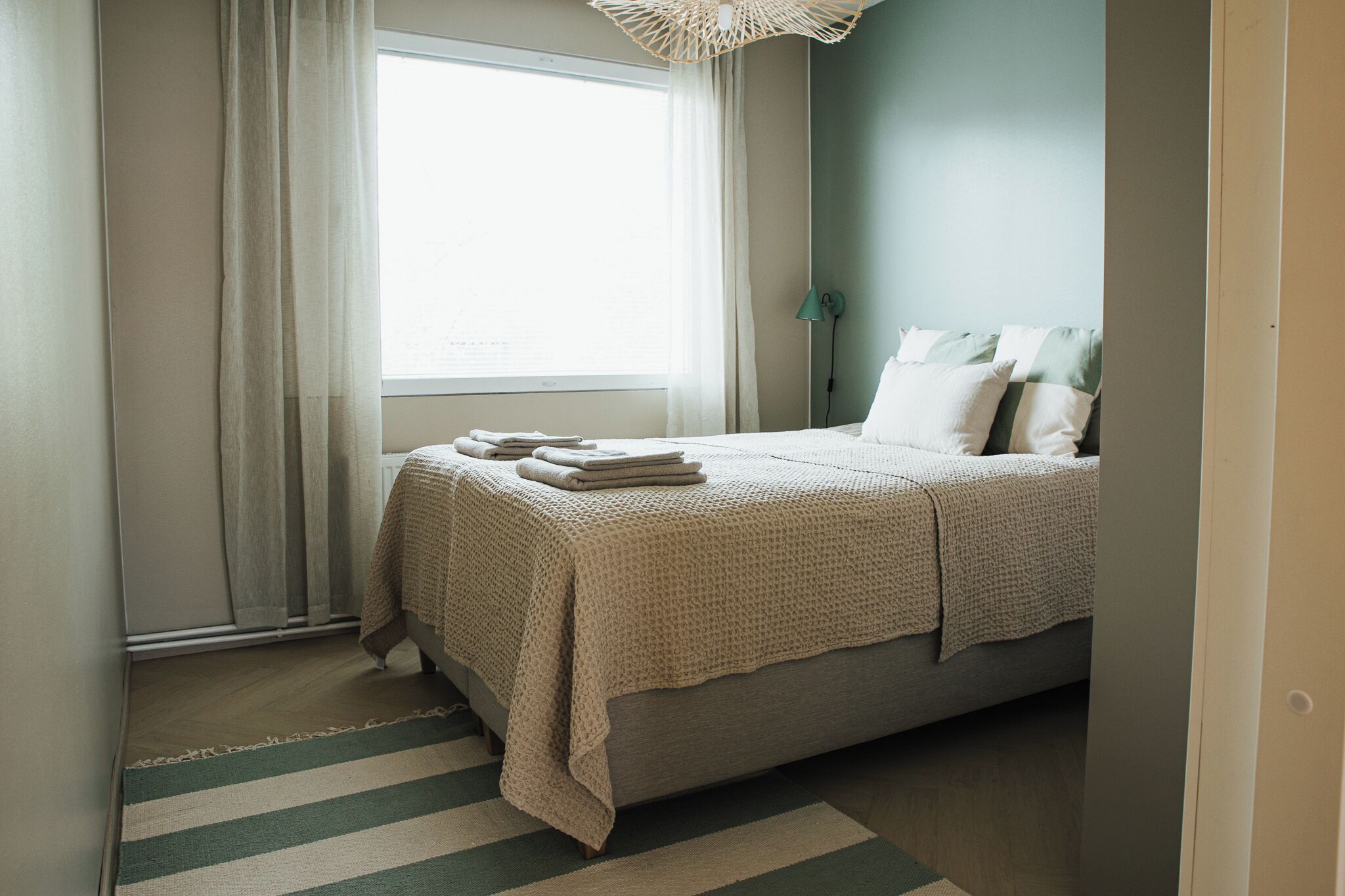 A bedroom with a bed, a window with curtains, a bedside lamp, and a striped rug.