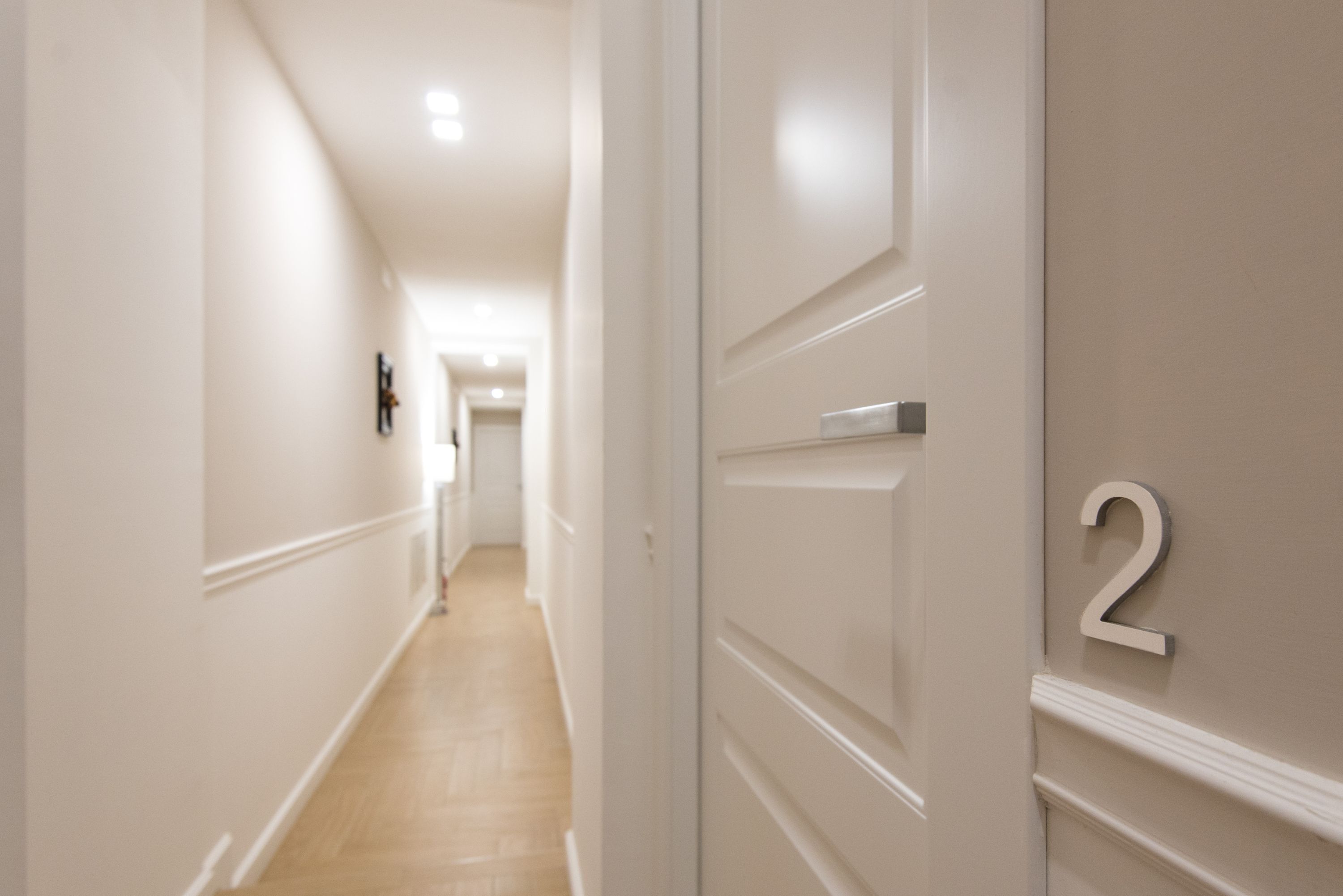 Un passadís amb sòl de fusta i parets blanques, que condueix a una habitació amb una porta que porta el número 2.