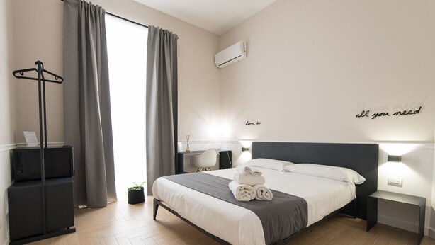 A modern hotel room with a bed, bedside tables, a desk, a chair, a wardrobe, and a large window with curtains. - Five Boutique Rooms - Camera 2 (Naples)
