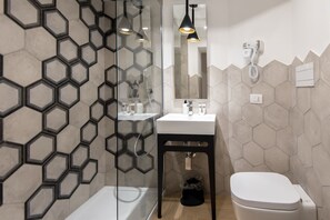 Ein modernes Badezimmer mit sechseckischem Fliesenmuster, einem weißen Waschbecken, einer schwarzen Kommode und einer weißen Toilette sowie einer Duschkabine aus Glas.
