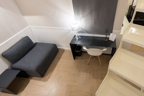 Ein gemütlicher Raum mit grauem Sofa, kleinem Schreibtisch mit Lampe, weißem Stuhl und weißem Schrank.