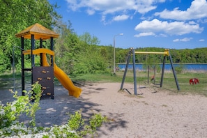 A playground with a slide and swings, surrounded by trees and a body of water. - Modern vacation home in Korswandt with a garden, fireplace, and a view of the lake nearby (Korswandt)