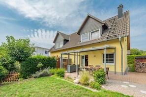 A two-story house with a covered patio, a garden, and a visible chimney. - Modern vacation home in Korswandt with a garden, fireplace, and a view of the lake nearby (Korswandt)