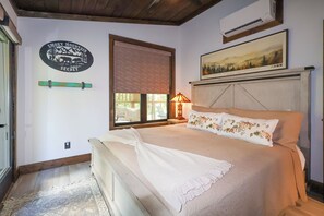 A bedroom with a bed, a window with blinds, a wall-mounted air conditioner, and a painting above the bed. - Gorgeous new 1 bedroom with king bed, outdoor firepit, hot tub, lots of light! (Sevierville)