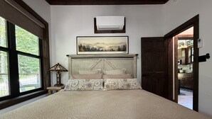 Una camera da letto con un letto grande, una testiera con un quadro, una finestra vista alberi e una porta che dà su una cucina.