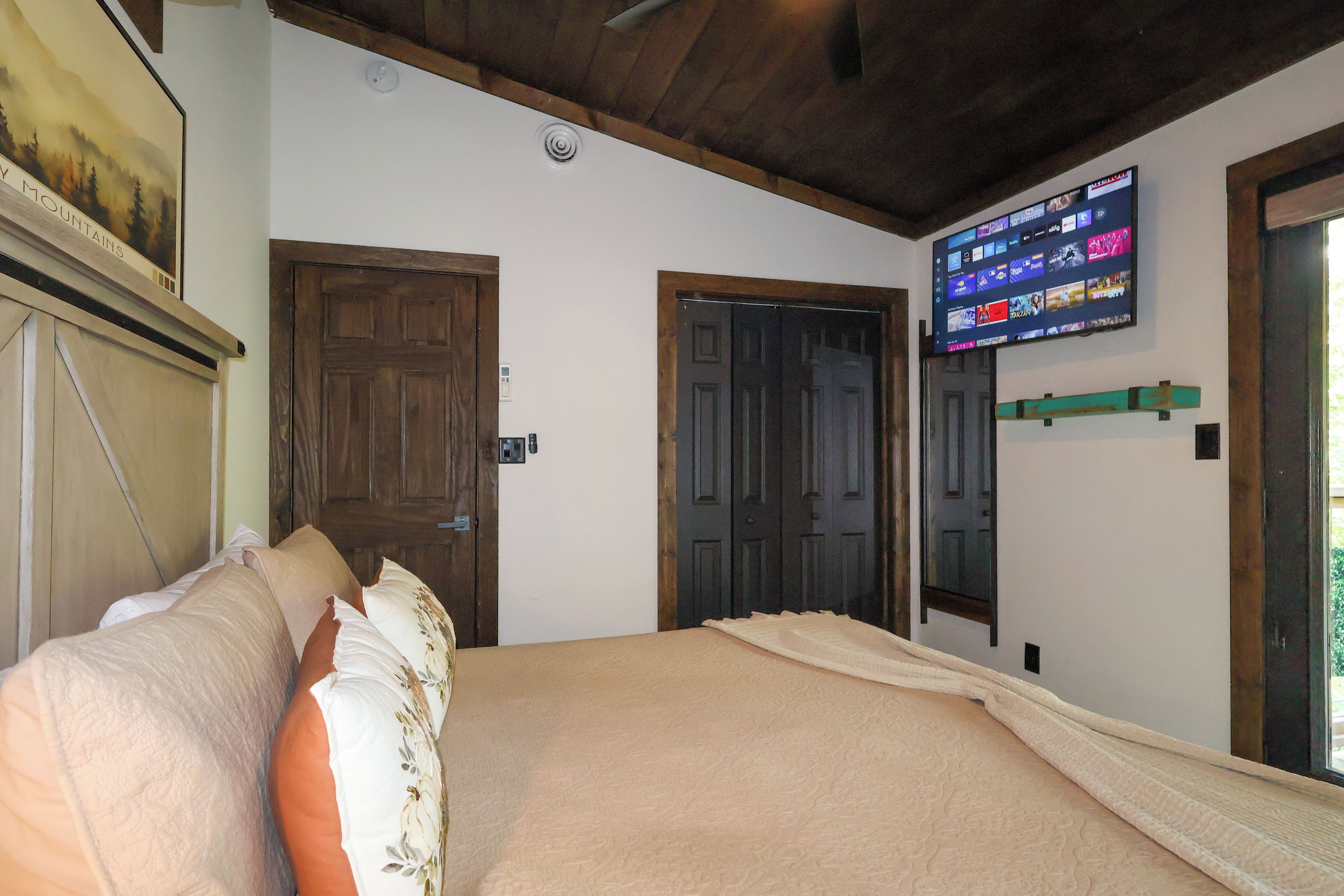 A bedroom with a bed, two doors, a mounted TV, and a painting on the wall.