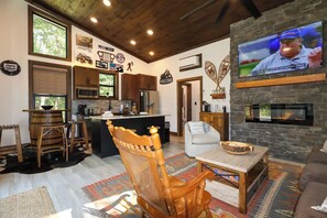A cozy living room with a fireplace, a television mounted on the wall, and a kitchen area with wooden cabinets and a bar stool. - Gorgeous new 1 bedroom with king bed, outdoor firepit, hot tub, lots of light! (Sevierville)