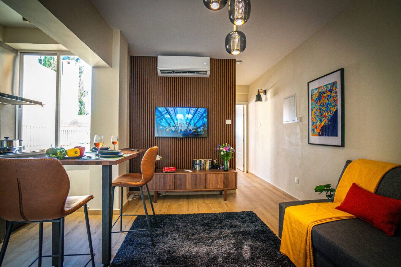 A modern living room with a sofa, a TV, a dining area with a table and chairs, and a kitchen area with a window.