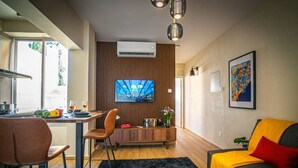 Una sala moderna con sofá, televisión, zona de comedor con mesa y sillas, y espacio de cocina con ventana.