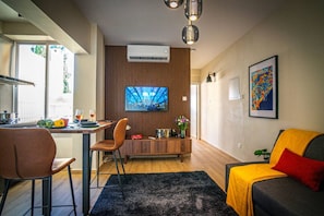 Ein modernes Wohnzimmer mit Sofa, Fernseher, Essbereich mit Tisch und Stühlen sowie Küchenzeile mit Fenster.