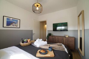 Une chambre d’hôtel comprenant un lit, une télévision, un tableau, une armoire en bois et un panier rempli d’objets divers.