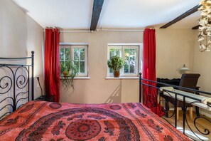 A room with a bed, two windows with curtains, a potted plant, and a desk with a lamp.