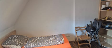 A small, single-bed room with a sloped ceiling, a wooden floor, and a built-in shelf with folded clothes.