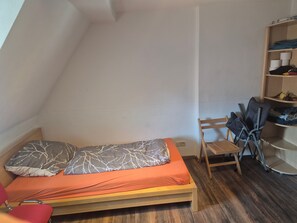 Una piccola camera con un letto singolo, soffitto spiovente, pavimento in legno e una mensola a incasso con vestiti piegati.