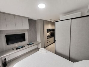 Moderni hotellihuone, jossa on taulutelevisio, yöpöytä, sohva ja vaatekaappi.