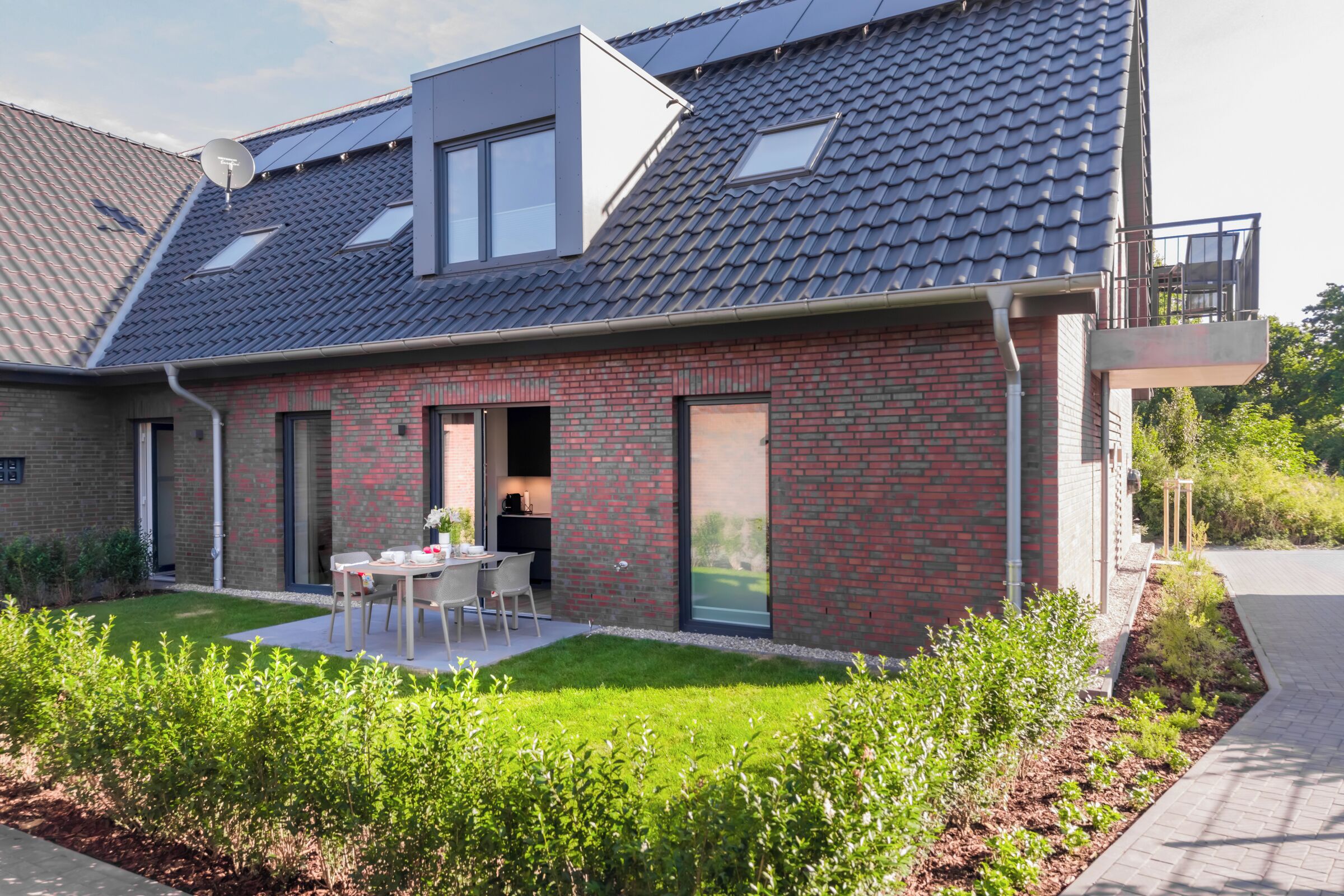 Ein modernes Haus mit Ziegelaußenwand, einer flachen Dachterrasse mit Solaranlage, einer sichtbaren Küche und einer kleinen Terrasse mit Tisch und Stühlen.