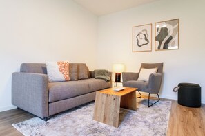 Una sala de estar moderna con un sofá gris, una mesa de centro de madera, una mesita de noche con una lámpara, un sillón gris y dos obras de arte abstracto en la pared.
