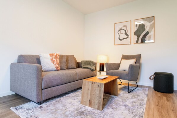 Een moderne woonkamer met een grijze bank, een houten salontafel, een bijzettafeltje met een lamp, een grijze fauteuil, en twee abstracte kunstwerken aan de muur.
