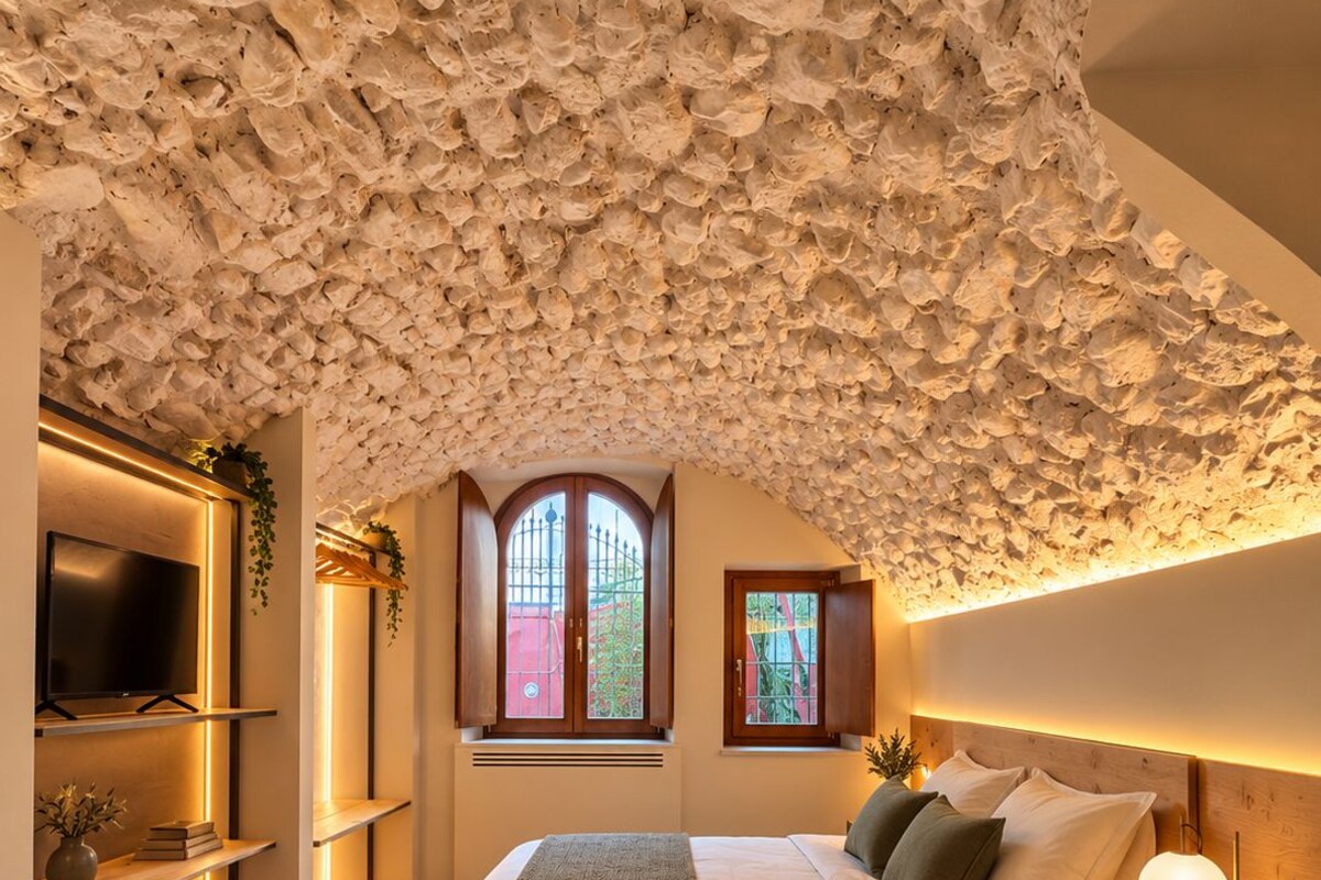 Un dormitorio con techo de piedra, una cama con almohadas, una mesita de noche con lámpara y un televisor en un estante.