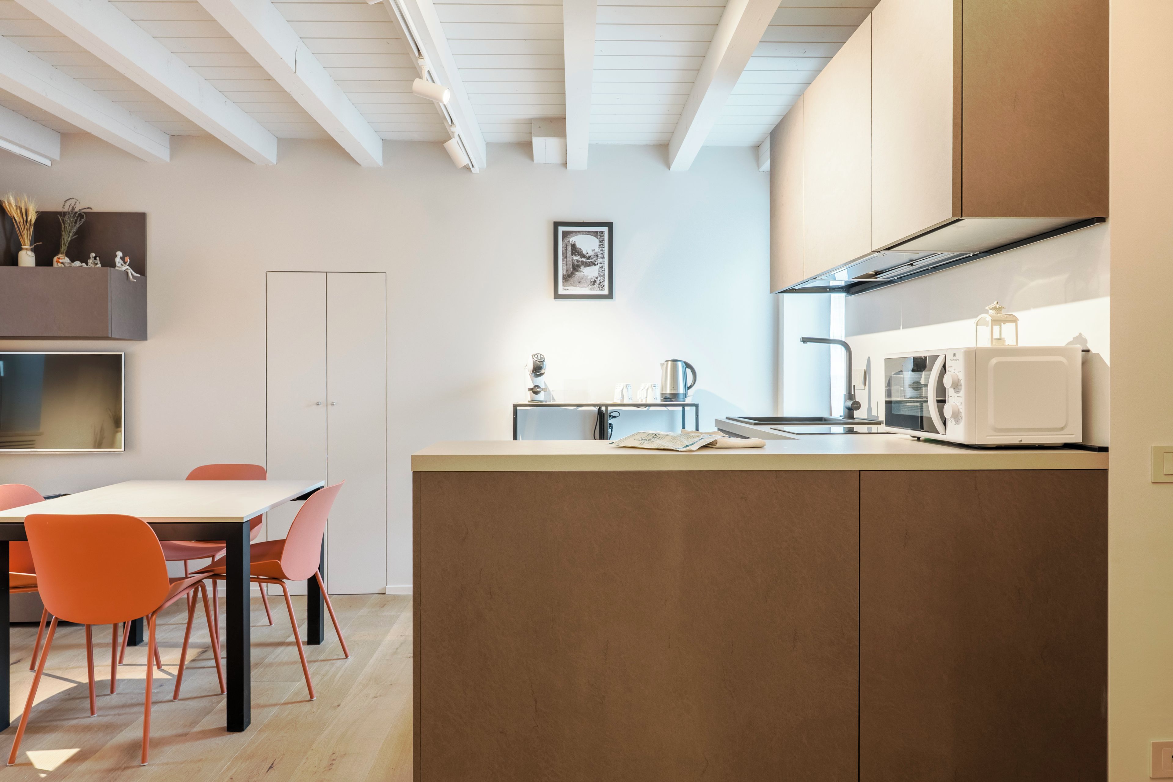 Een moderne keuken met een centraal eiland, een eetgedeelte met een tafel en stoelen, en een wit plafond met zichtbare balken.