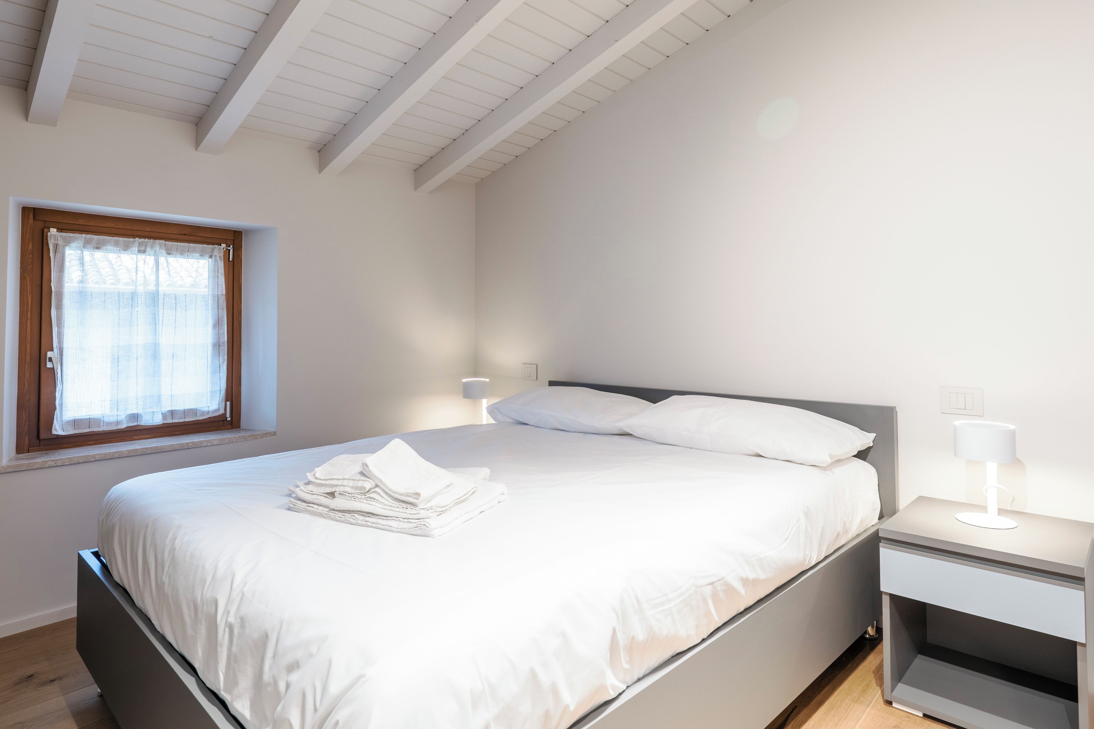 Un dormitorio con cama, mesita de noche, lámpara, ventana con cortinas y techo de madera.