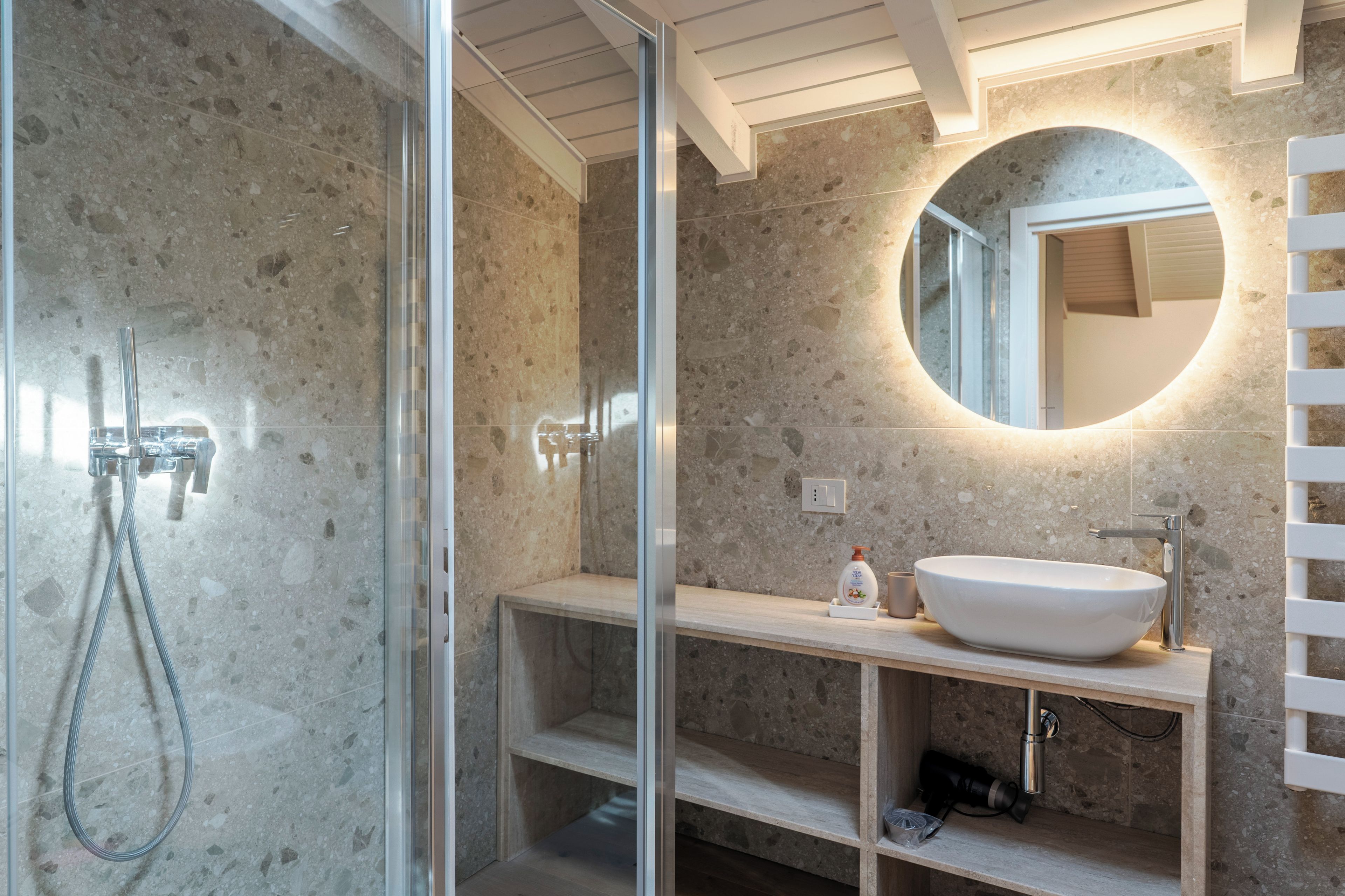 Ein modernes Badezimmer mit einer Duschkabine aus Glas, einer Wand mit Marmorplatten, einem runden Spiegel und einem weißen Waschbecken.