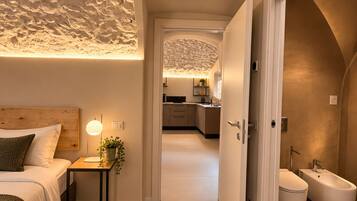 Un dormitorio moderno con techo texturizado, cabecera de madera, mesita de noche con lámpara, y vistas a la cocina y el baño a través de una puerta abierta.