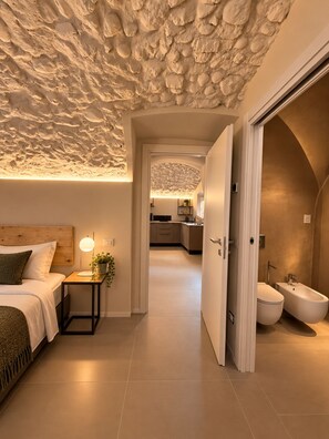 Une chambre moderne avec un plafond texturé, une tête de lit en bois, une table de chevet avec une lampe, et une vue sur la cuisine et la salle de bain grâce à une porte ouverte.