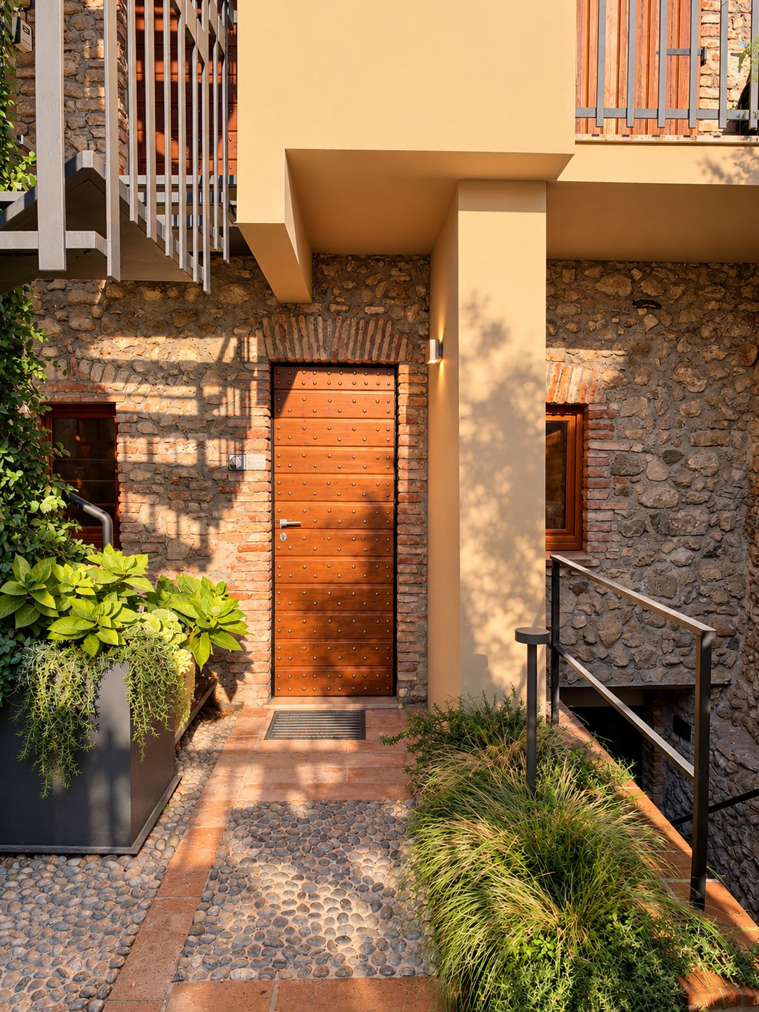 Fachada de piedra con puerta de madera, escalera de metal y maceta con plantas verdes.
