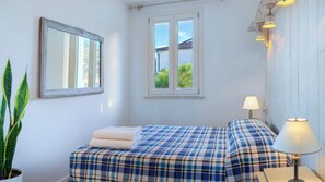Ein Zimmer mit einem Bett, einem Nachttisch mit Lampe, einem Fenster mit Blick auf Grünflächen und einem Spiegel an der Wand.