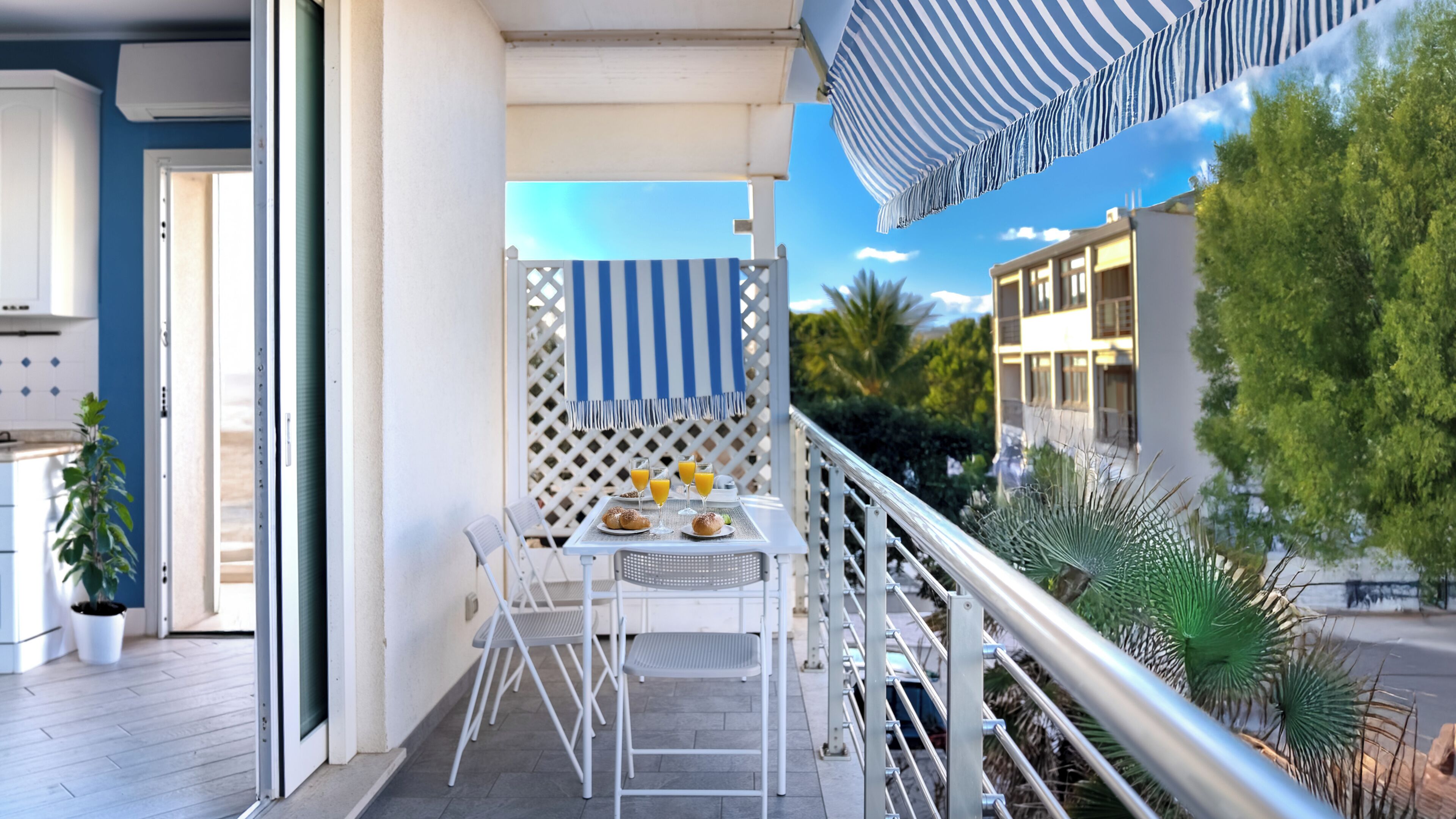 A balcony with a table set for two, a striped awning, and a view of a building and trees.